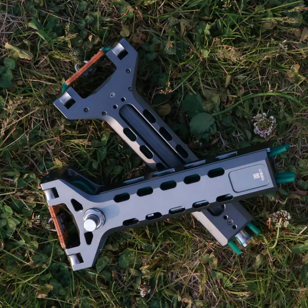Two heavy-duty AutoFling auto-loading slingshots resting on green grass after an outdoor field test, featuring a dark grey finish and high-tension bandsets.