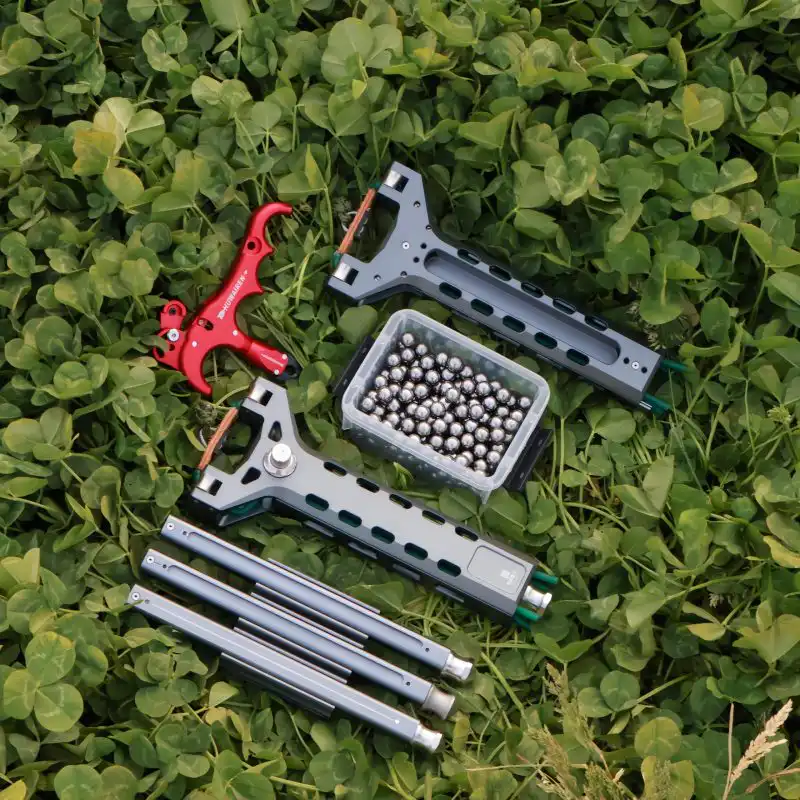A dynamic outdoor display of the AutoFling auto-loading slingshot Duo Kit on clover, featuring two dark grey CNC aluminum chassis, a red mechanical release aid, three speed dispensers, and a box of 8mm carbon steel ball bearings.